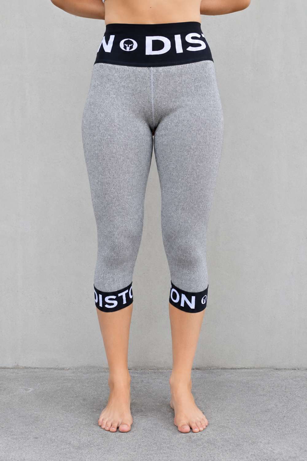 A person wearing DISTON cut resistant pants leggings with black elastic waistband and calf bands featuring white text, shown from the back against a plain gray wall.