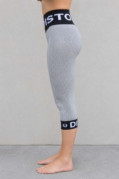 Side view of a person wearing DISTON cut proof pants leggings with black elastic waistband and ankle cuffs featuring white branding text, standing barefoot against a light gray wall.