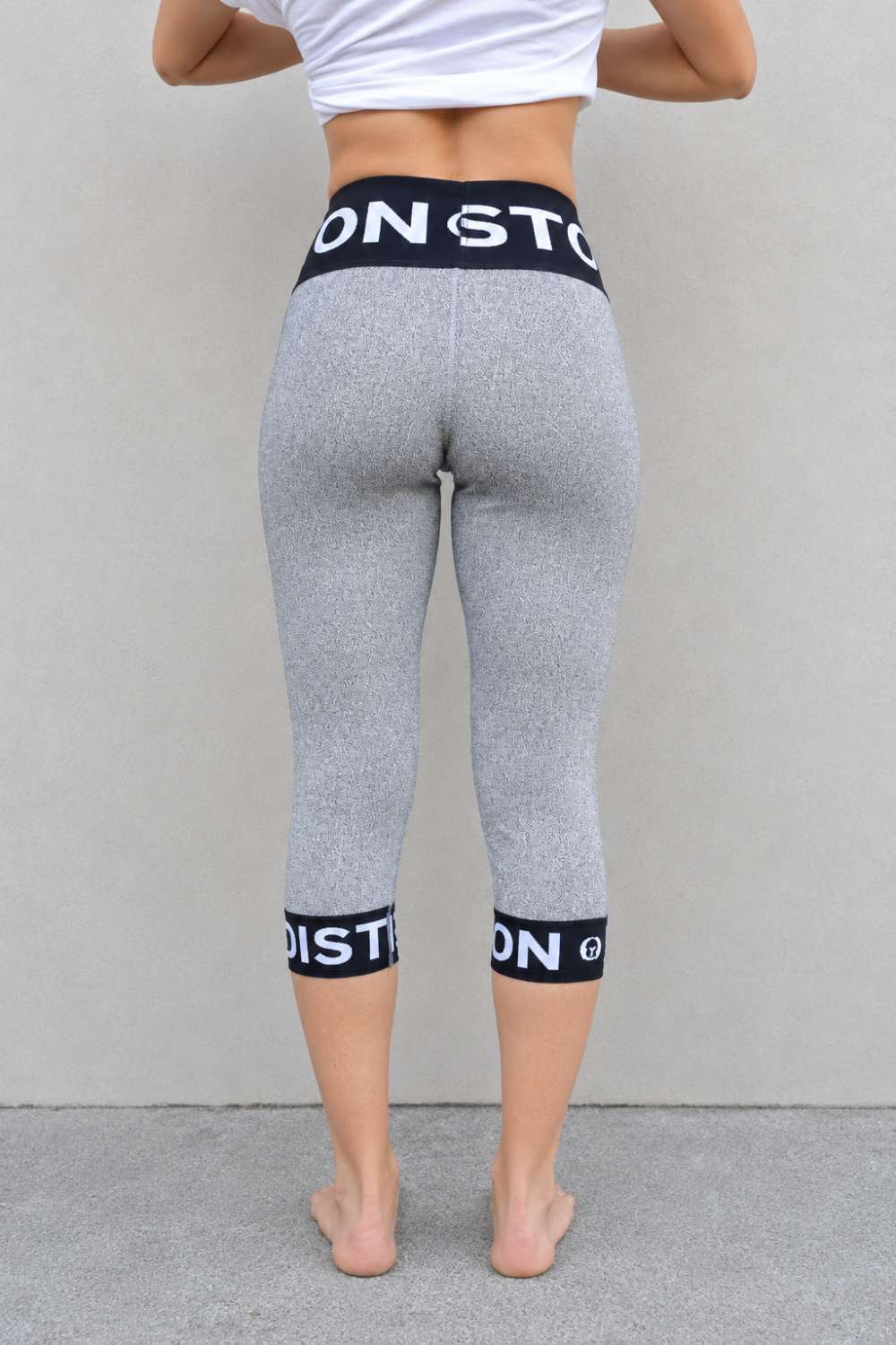 A person shown from the back wearing heather gray capri leggings with black elastic waistband and calf bands featuring white text, standing against a plain light gray wall.