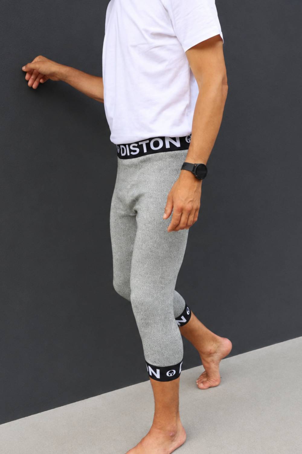 A man wearing heather gray cropped leggings with black elastic waist and ankle bands featuring white branding, paired with a white t-shirt, standing against a dark gray wall.