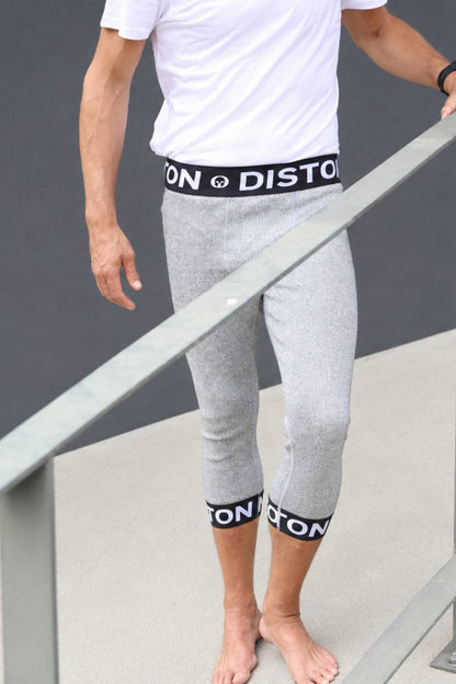 A man wearing light gray heathered cropped capri pants with black elastic waist and ankle bands featuring white text, standing barefoot on a concrete surface next to a metal railing.