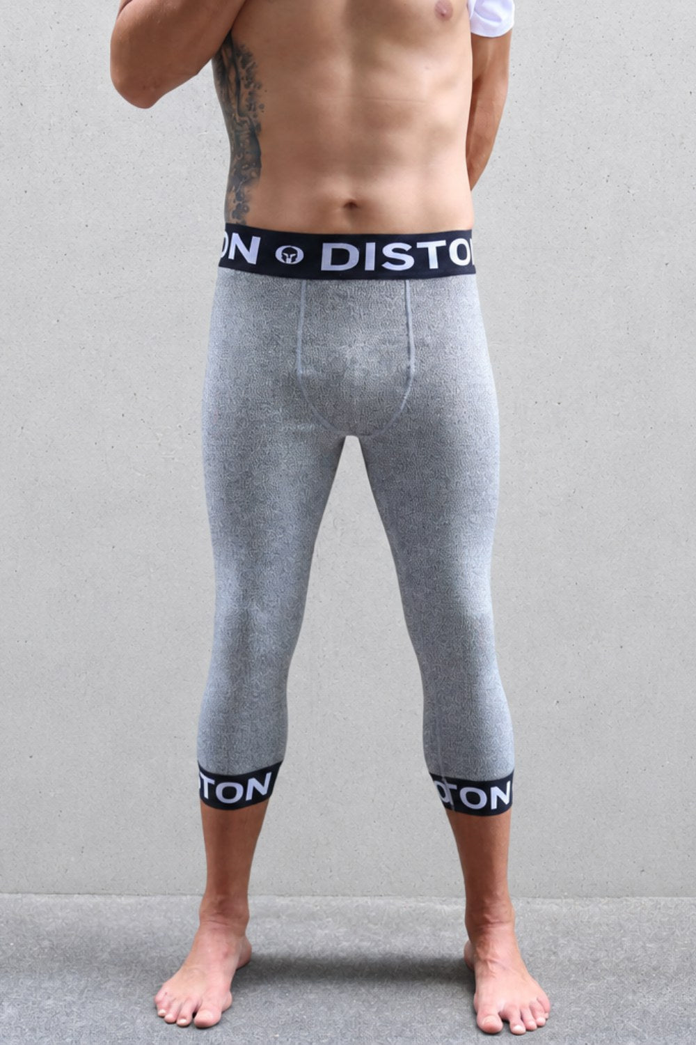A man ski racer wearing heather gray full-length compression leggings with black elastic cuffs and a matching waistband featuring the white 'DISTON' brand logo, standing against a gray wall.