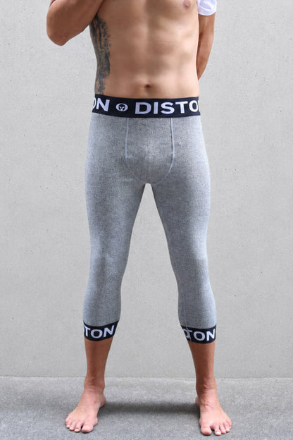 A man ski racer wearing heather gray full-length compression leggings with black elastic cuffs and a matching waistband featuring the white 'DISTON' brand logo, standing against a gray wall.