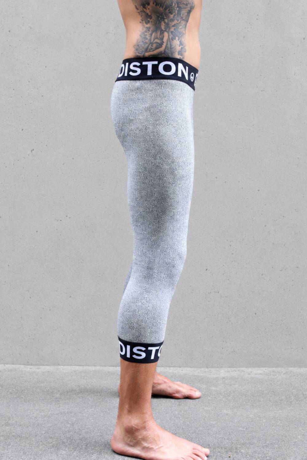 A side view of a person's legs wearing heather gray compression cut resistant pants with black and white Diston brand bands at the top and bottom, standing against a gray concrete wall.