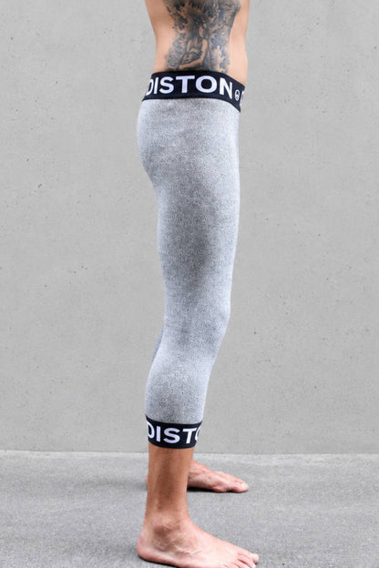 A side view of a person's legs wearing heather gray compression cut resistant pants with black and white Diston brand bands at the top and bottom, standing against a gray concrete wall.