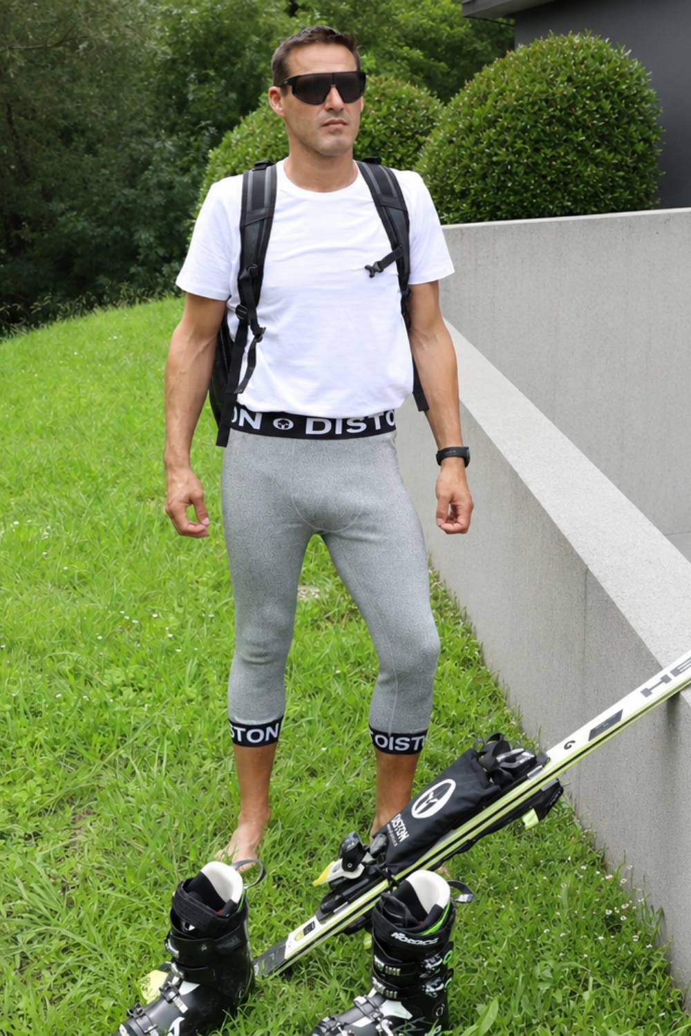 A man wearing a white t-shirt, gray capri leggings with black Distron branding, black ski boots, and a black backpack stands on a grassy slope next to a concrete wall with skis in the foreground.
